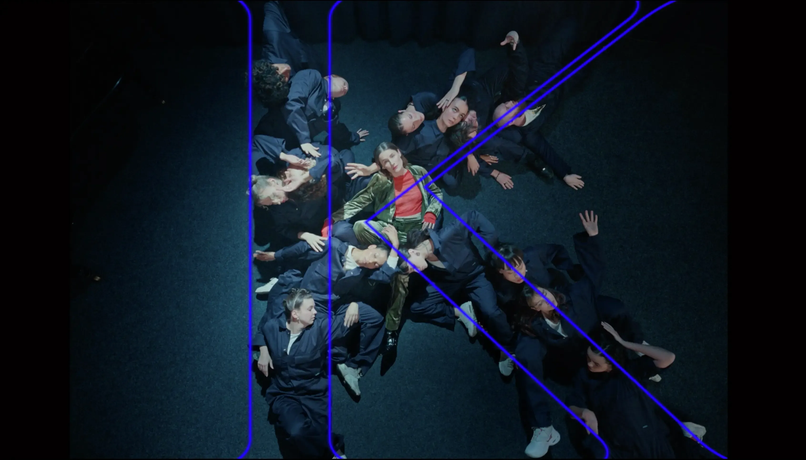 Lidé ležící na zemi jeden přes druhého ve tvaru písmene K.